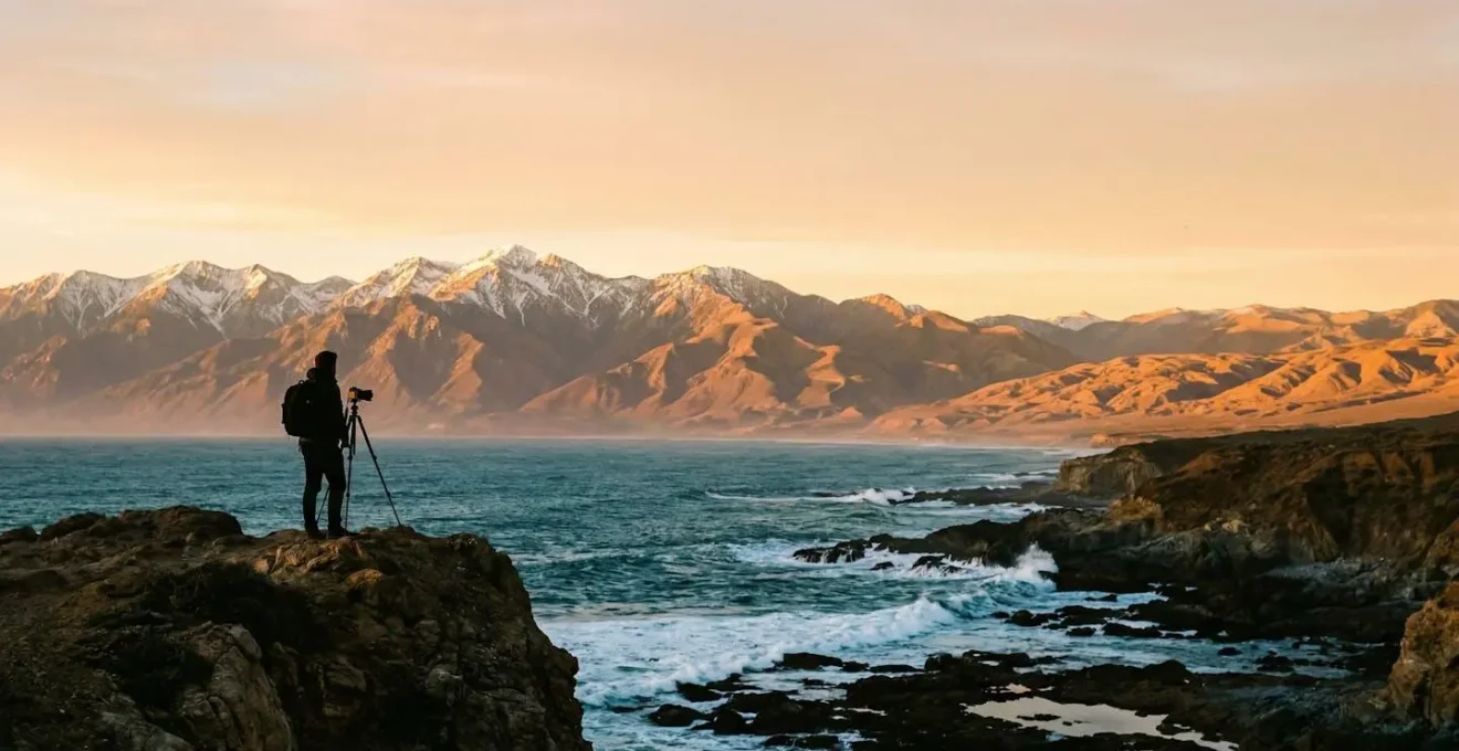 Photographer silhouetted against dramatic transitional landscape where coastal cliffs meet mountain foothills with desert visible in distance